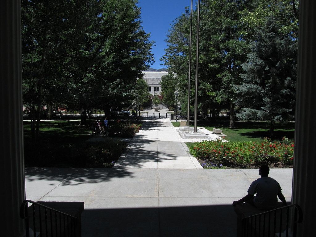 View from a shaded portico down a tree-lined walkway with flowerbeds and flagpoles toward a stone building