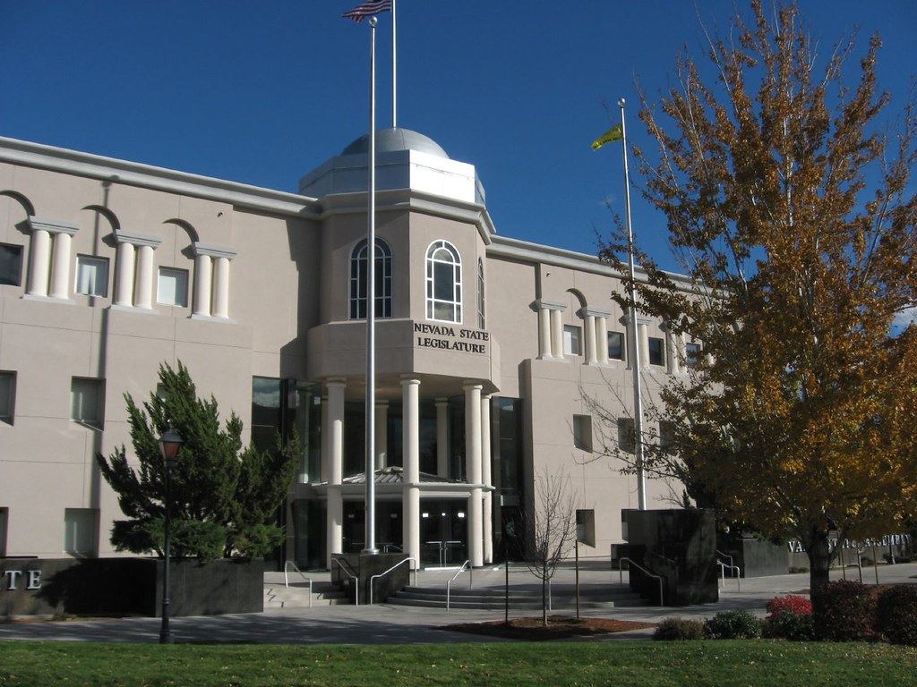 Exterior of the Nevada State Legislature building with flagpoles, columns and an autumn tree under a blue sky