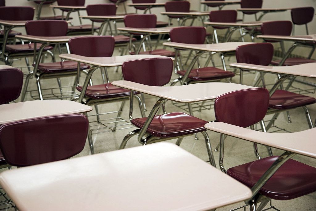 Rows of empty classroom desk-chairs with maroon seats and beige writing surfaces