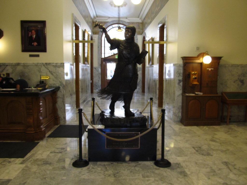 Bronze statue on a pedestal behind rope barriers in a marble hallway with wood furnishings and an arched doorway