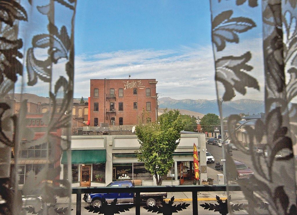 View through lace curtains of a small-town street with storefronts, a brick building and mountains in the distance