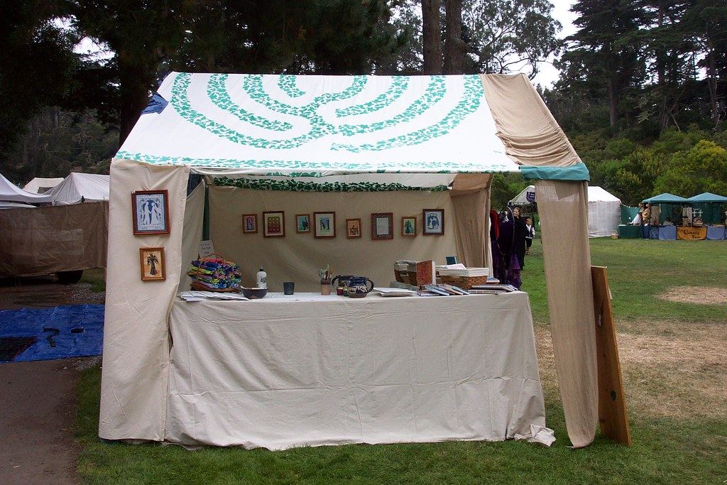 A canvas craft booth with framed artwork and a draped table, with other fair tents on a grassy field behind it