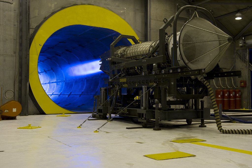 A jet engine mounted on a test stand fires a blue exhaust flame into a large yellow-rimmed tunnel indoors