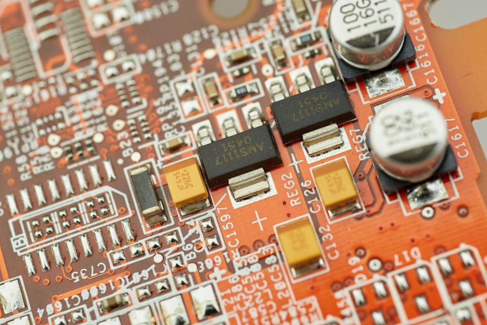 Close-up of an orange circuit board with black voltage regulators, yellow capacitors and silver capacitors
