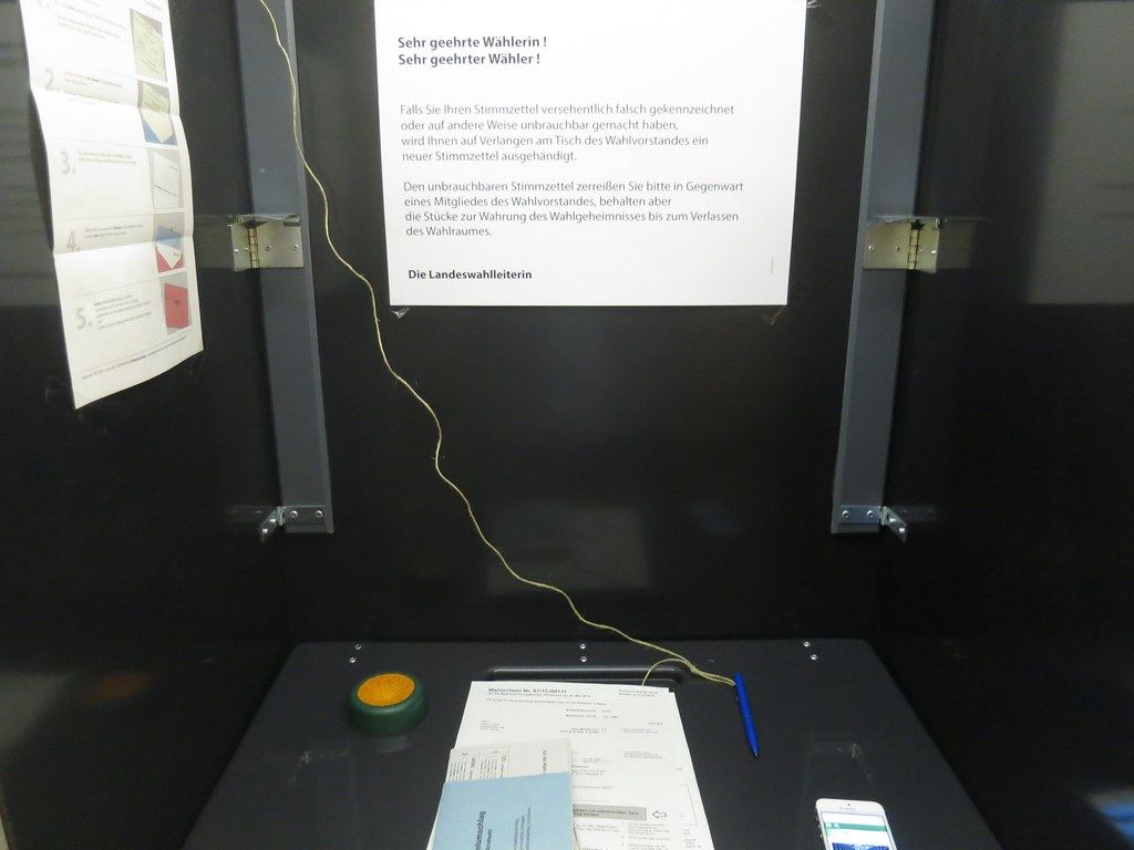 Inside a dark voting booth with ballot papers, a pen on a string, a smartphone and printed instructions on the wall