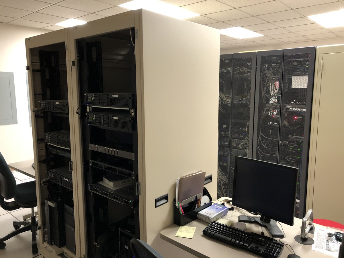 Server racks with blinking equipment and cables beside a desk with a monitor and keyboard in an office room