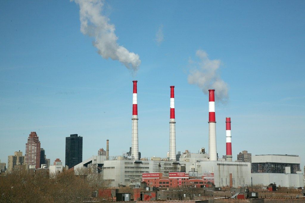 A power plant with four red-and-white striped smokestacks releasing white plumes against a blue sky beside city buildings