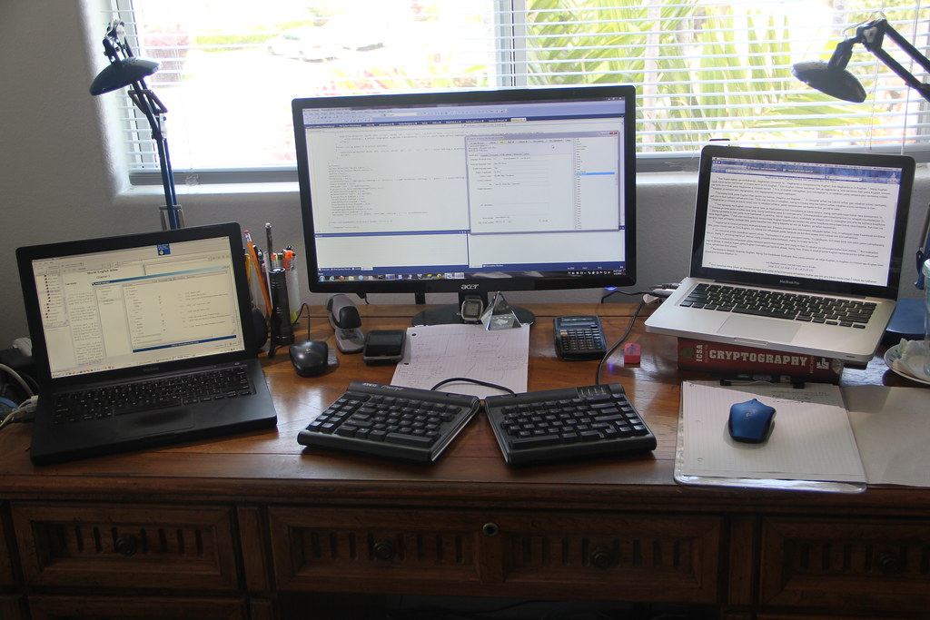 A wooden desk with two laptops, a monitor, a split keyboard, desk lamps and a mouse in front of a window