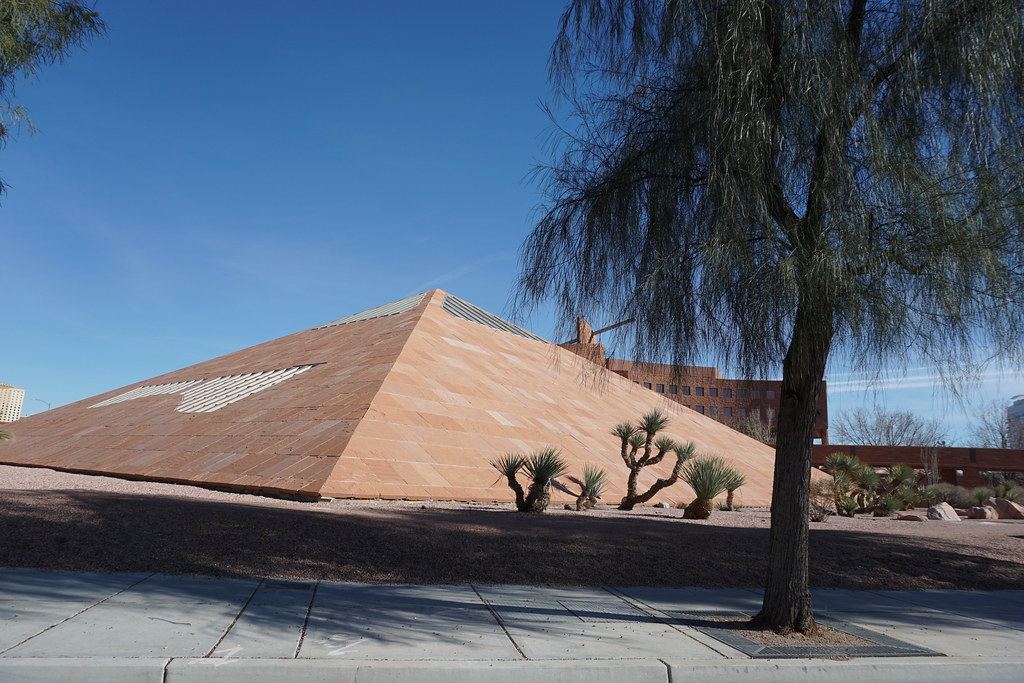 A sandstone pyramid-shaped structure with desert plants and a tree beside a sidewalk under a clear blue sky
