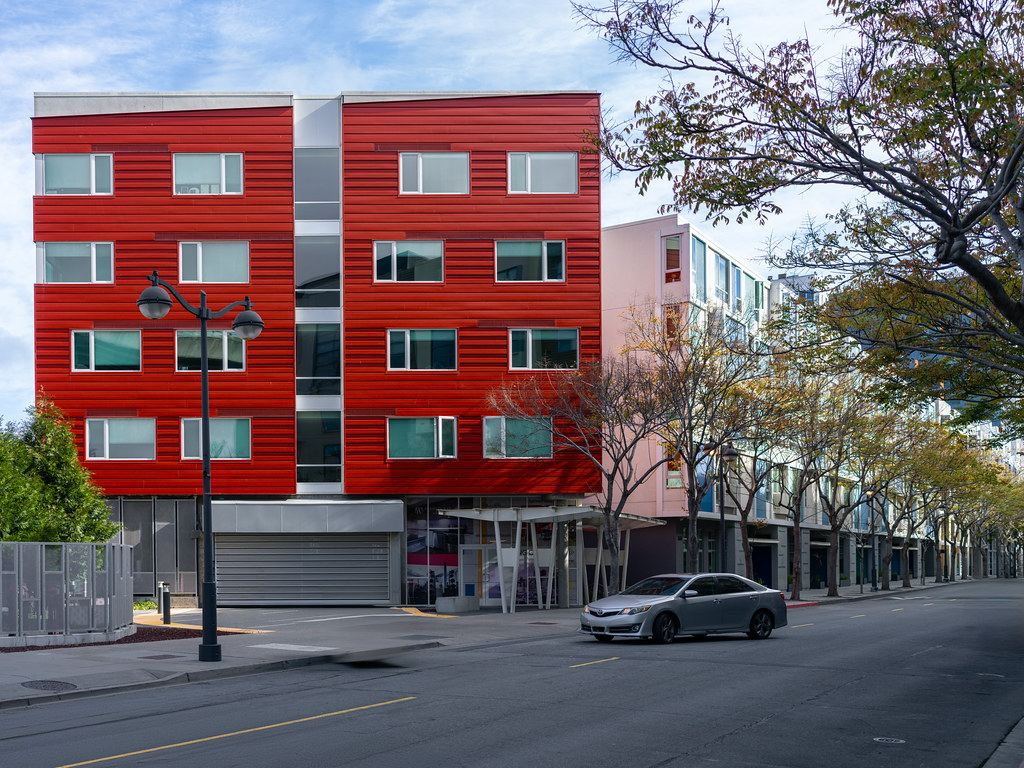 A red four-story apartment building beside a tree-lined street with a silver car driving past