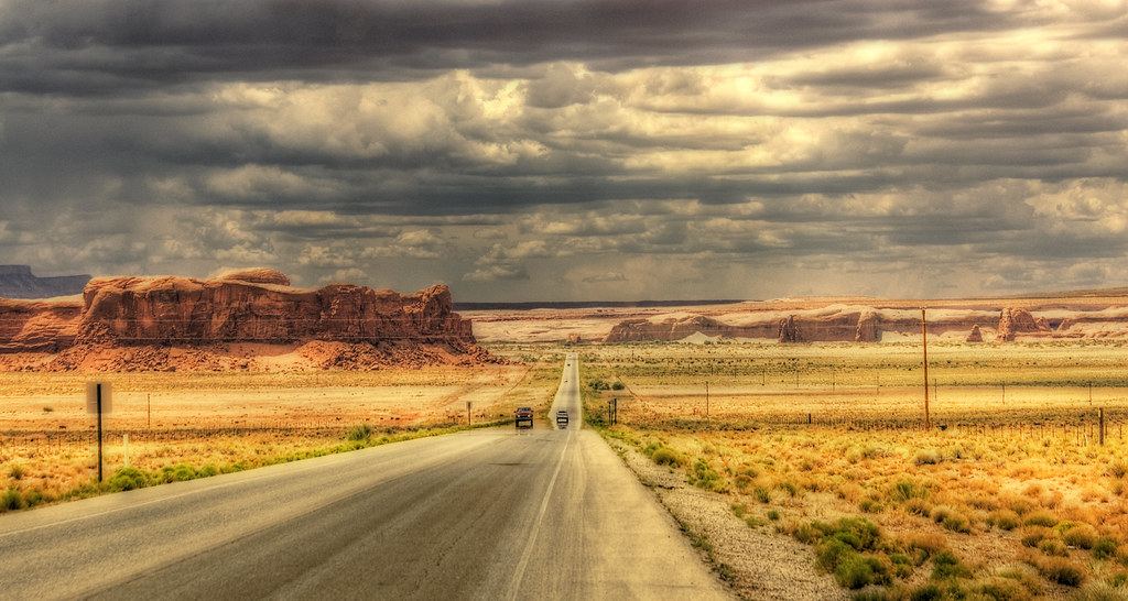 A straight desert highway runs past red rock buttes under dark, heavy storm clouds