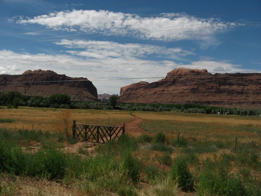 Red sandstone cliffs rise behind a grassy field with a wooden gate and dirt path under a blue, cloudy sky