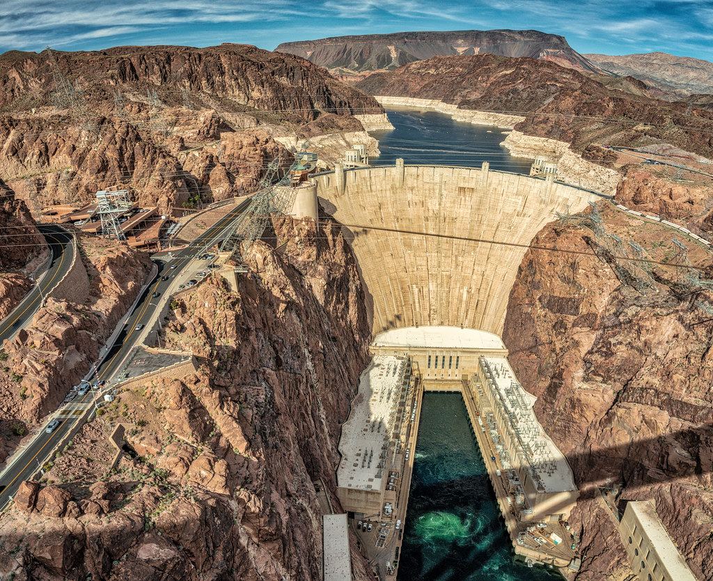 Aerial view of a curved concrete dam in a rocky canyon, with a reservoir behind it and a winding highway beside it