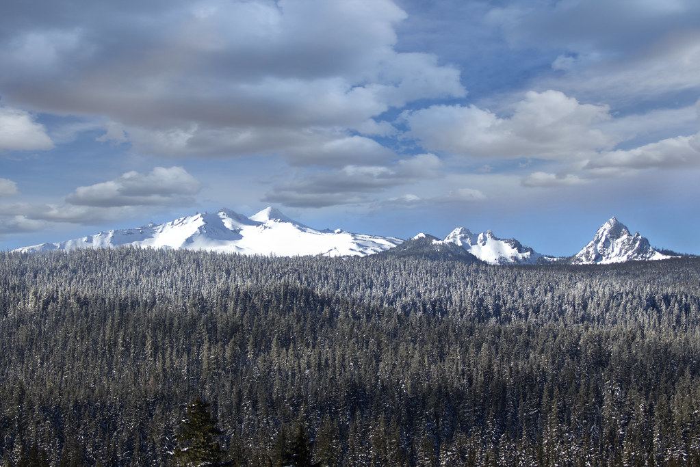 Snow-capped mountain peaks rise above a vast snow-dusted conifer forest under a cloudy blue sky