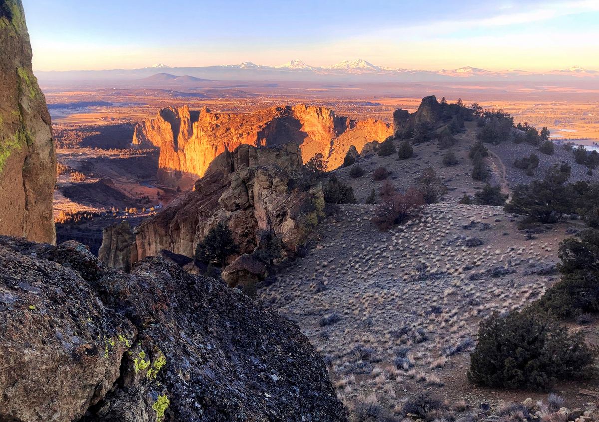 Sunlit rock cliffs and a sagebrush slope with snow-capped mountains on the horizon at dusk