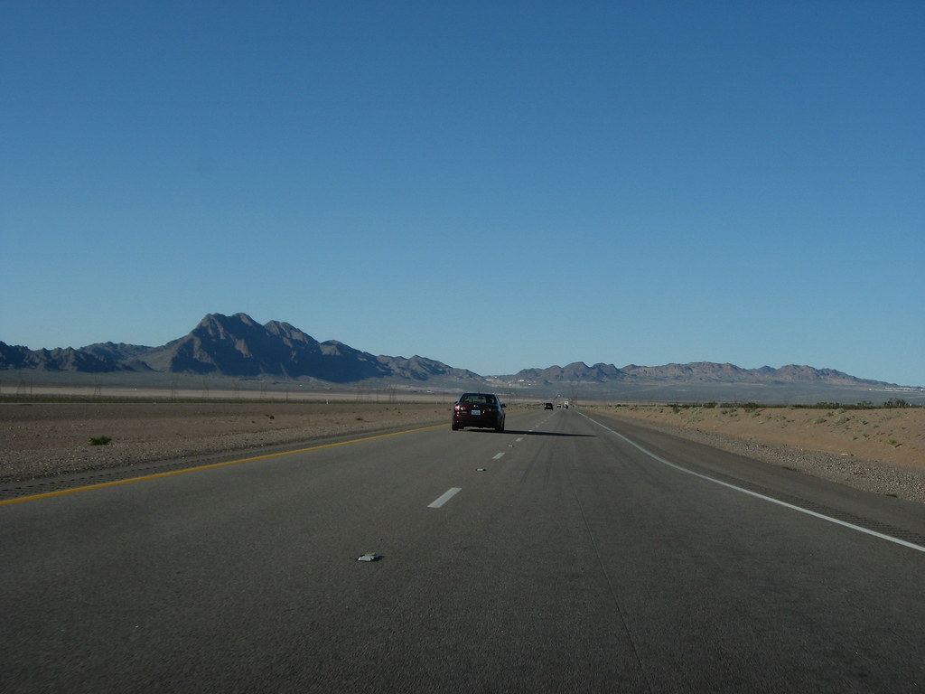 A car drives along a multi-lane desert highway toward rugged mountains under a clear blue sky