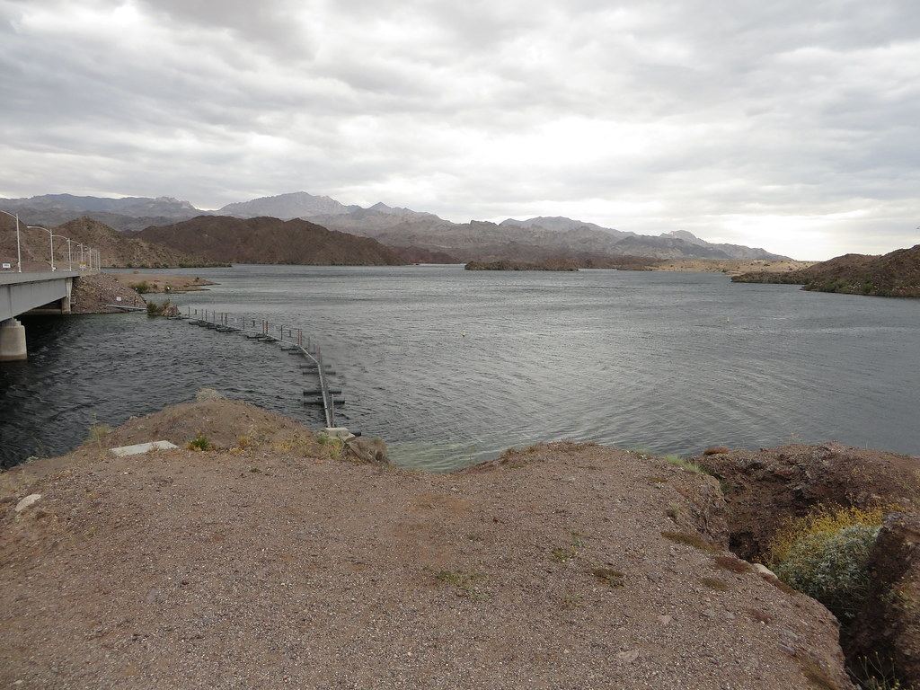 A desert lake under cloudy skies, with a bridge on the left, a floating barrier and rocky mountains beyond