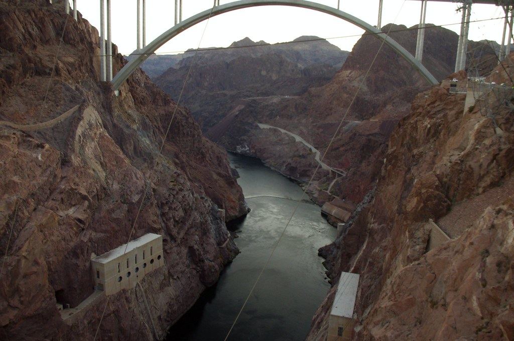 A river winds through a deep rocky canyon beneath a large concrete arch bridge, with small buildings on the cliffs