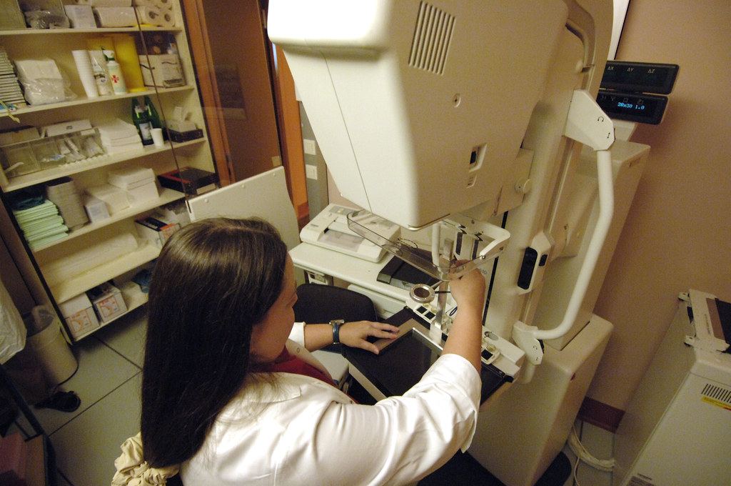 A technician in a white coat adjusts a mammography machine in a clinic room with shelves of supplies