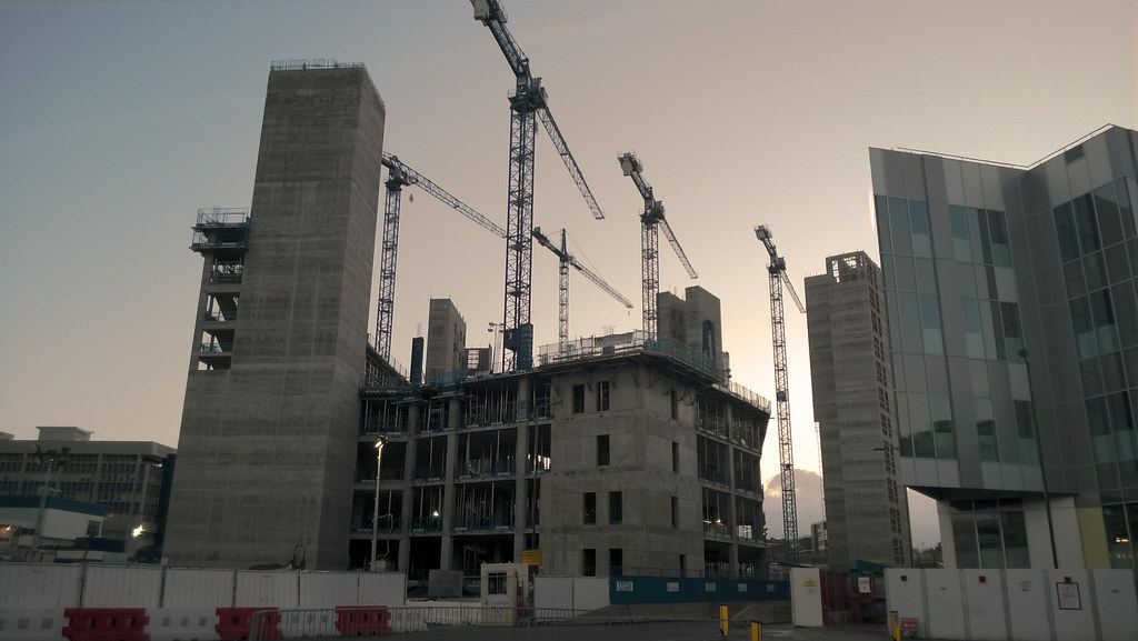 Several tower cranes rise over a partly built concrete building and core towers at dusk behind site fencing