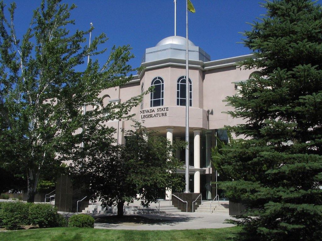 Domed Nevada State Legislature building with columns and front steps, framed by green trees under a blue sky