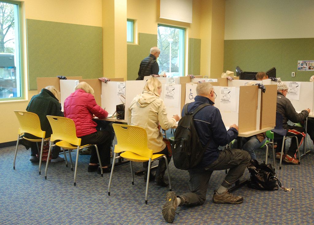 People seated and kneeling at cardboard voting booths in a room with yellow chairs and large windows