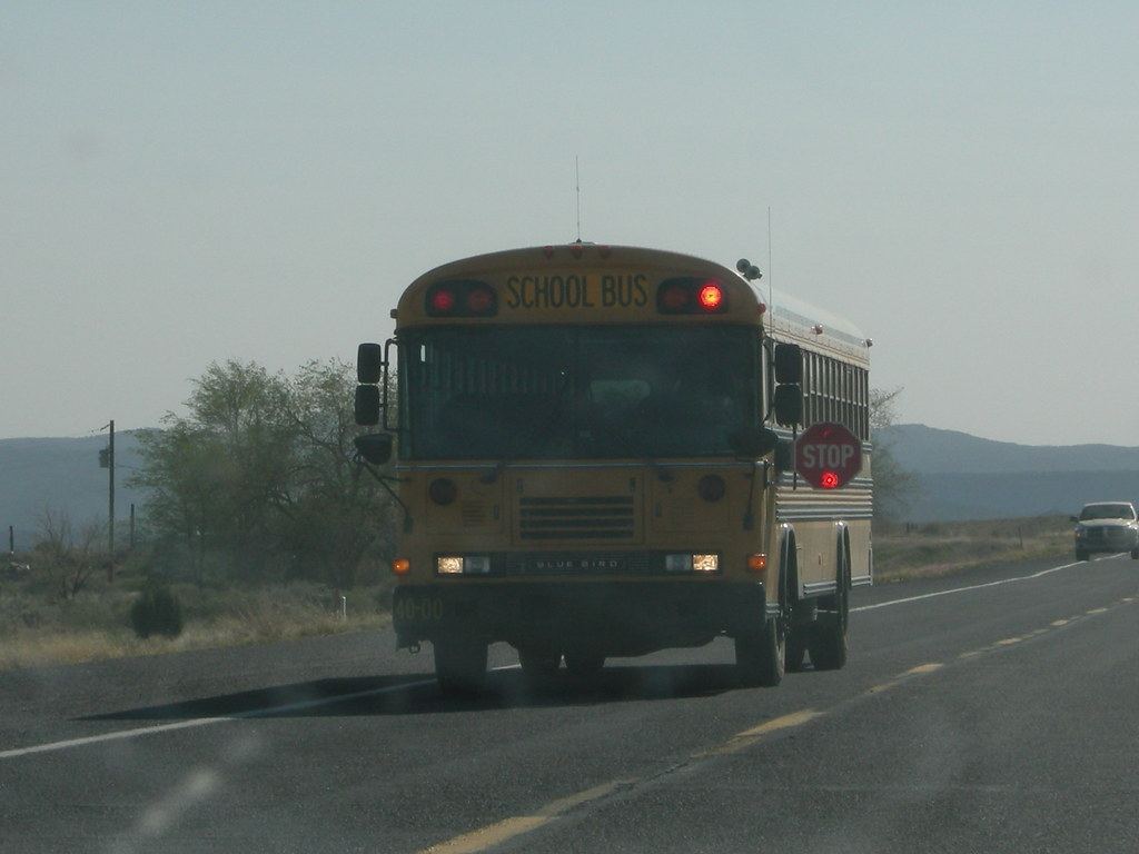 A yellow school bus with red warning lights on drives along a two-lane desert highway with hills behind