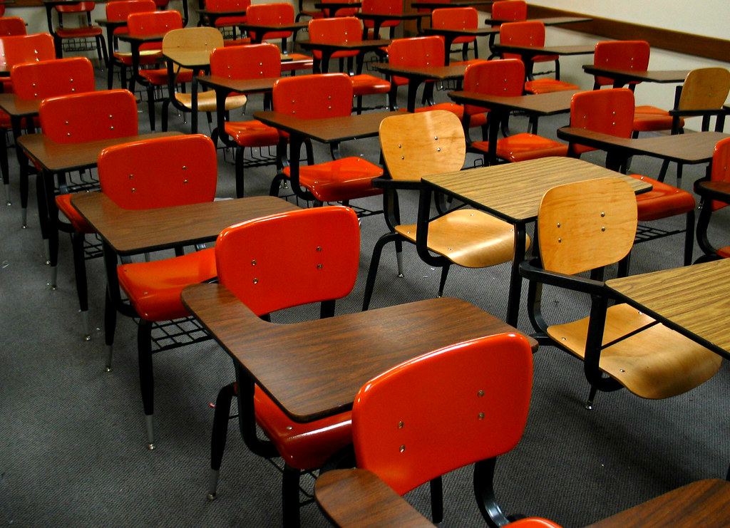 Rows of empty classroom desks with orange and wooden chairs on a gray carpet