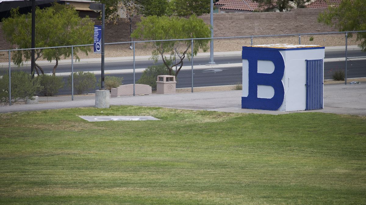 Grass athletic field beside a small white shed painted with a large blue letter B, behind a chain-link fence
