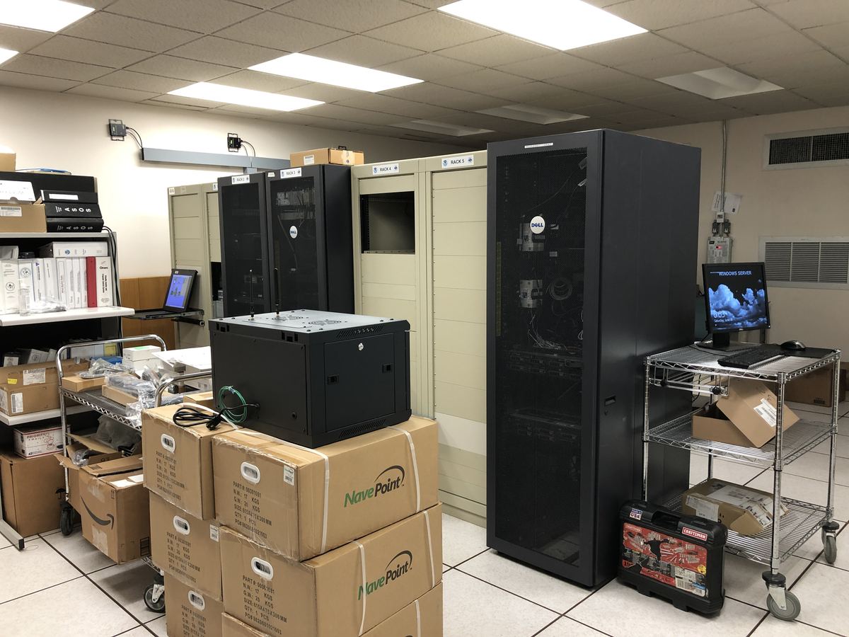 A server room with black and beige equipment racks, stacked cardboard boxes and a monitor on a wire cart
