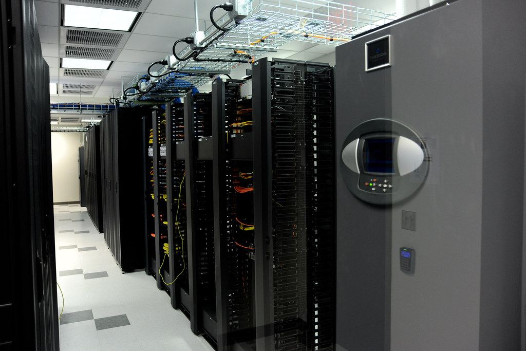 A row of black server racks with cabling beside a gray cooling unit in a brightly lit data center aisle