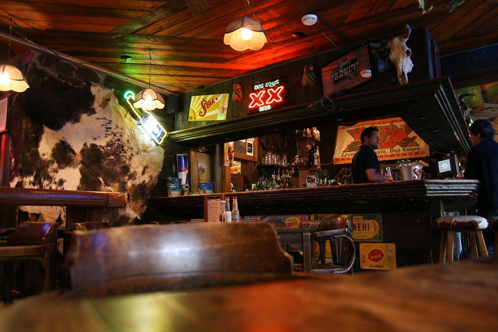 Dimly lit wooden bar with neon beer signs, a cowhide on the wall and empty stools in the foreground