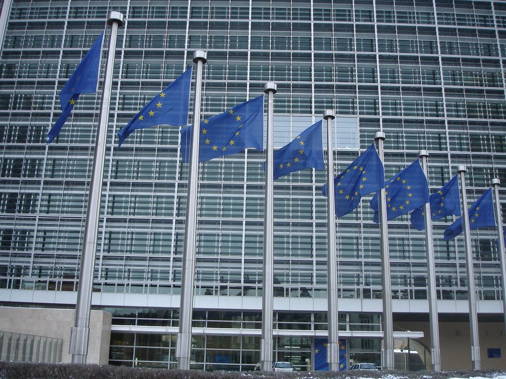 A row of European Union flags on tall poles in front of a large glass office building