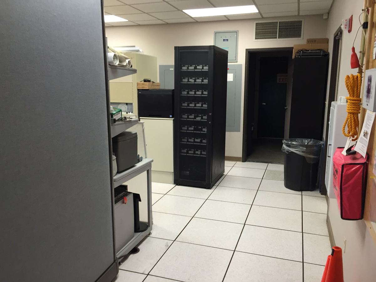 A small server room with a black equipment rack, shelves of supplies and a white raised-tile floor