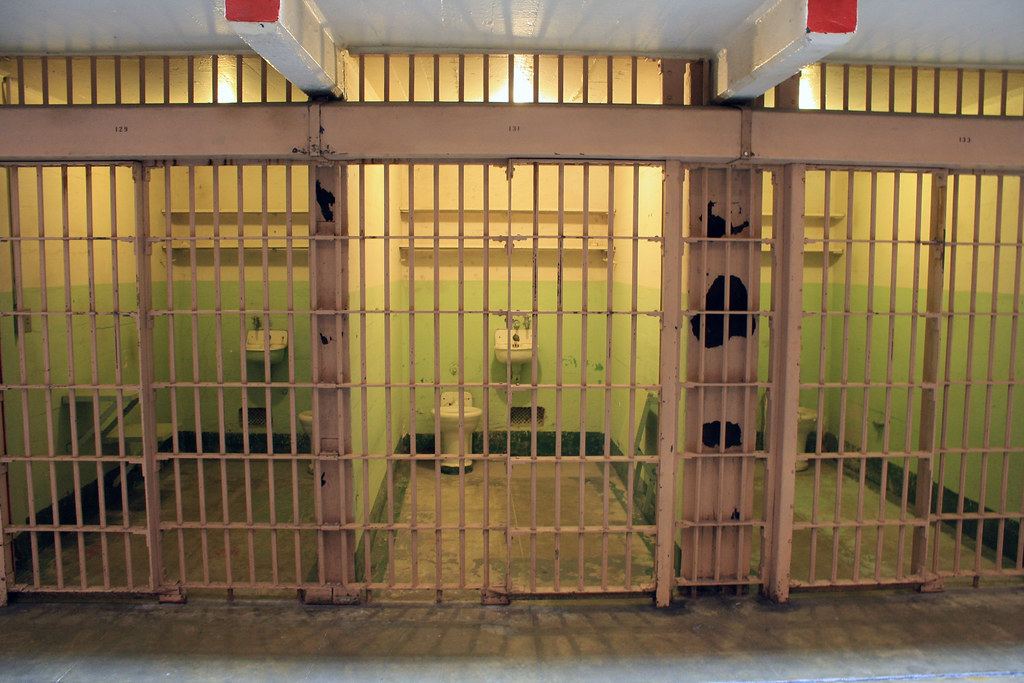 A row of empty prison cells behind barred doors, each with a sink and toilet against green-painted walls