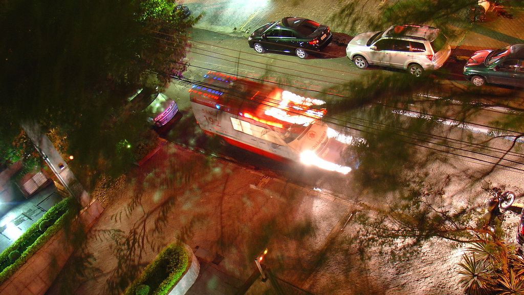 Overhead night view of an ambulance with flashing lights speeding past parked cars on a tree-lined street