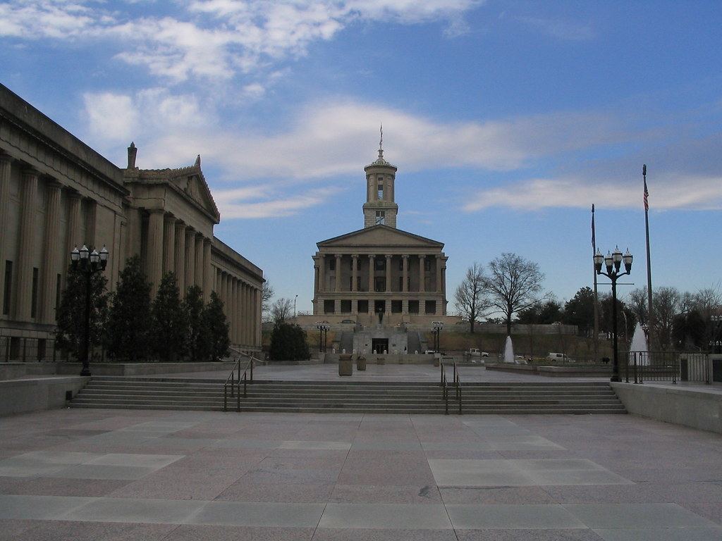 A columned capitol building with a tall cupola stands beyond a stone plaza and steps under a partly cloudy blue sky