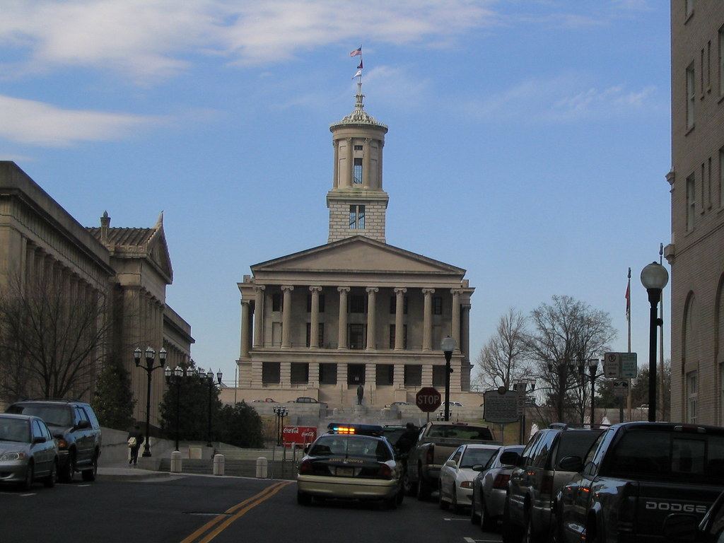 A columned stone capitol building with a tall tower at the end of a street lined with parked cars