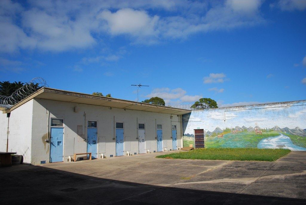 A low concrete cell block with blue doors and razor wire on the roof faces a yard beside a mural-painted wall