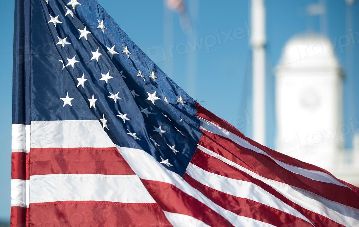 An American flag waves against a blue sky, with a blurred white tower in the background