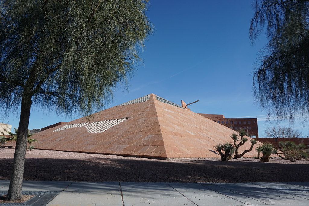 A sandstone-clad pyramid building with desert plants and trees beside a sidewalk under a clear blue sky