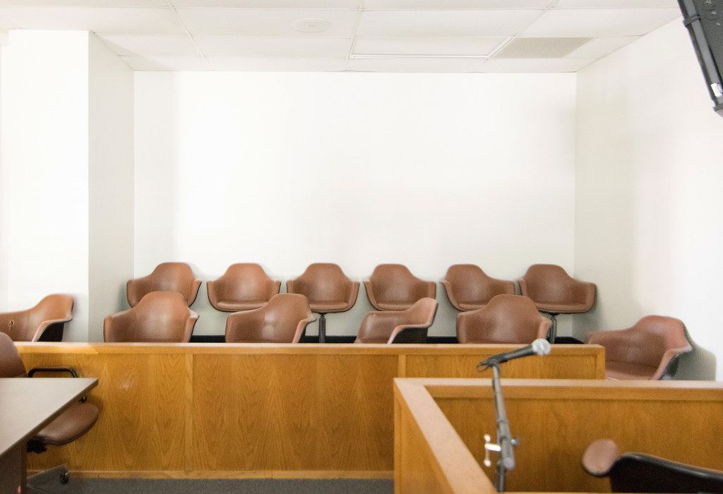 Empty courtroom jury box with brown chairs behind a wooden rail, and a witness stand microphone in the foreground