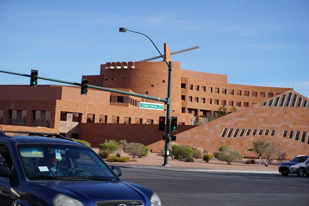 Red sandstone government building with a round tower behind a street intersection with traffic lights and a passing SUV
