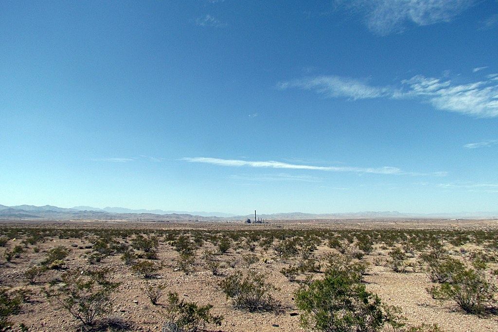 Flat desert scrubland under a blue sky with distant mountains and a lone industrial smokestack on the horizon