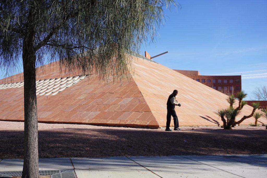 A red sandstone pyramid structure beside a brick building, with desert plants and a man walking past