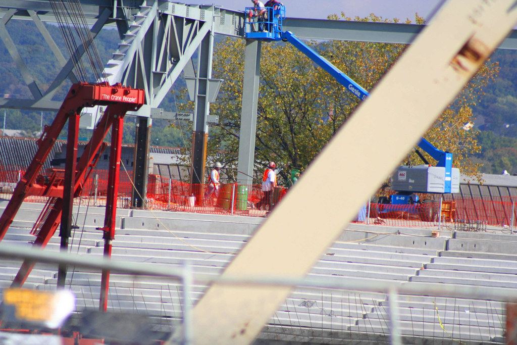 Workers and a blue boom lift among steel beams above concrete seating tiers at a stadium construction site