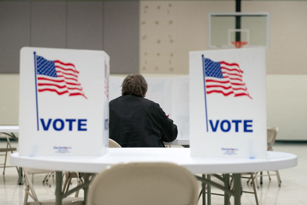 A person seated from behind at a voting booth between two white VOTE privacy screens with flags in a gym