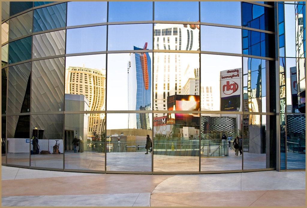Curved glass building facade reflecting high-rise towers and a plaza under a blue sky