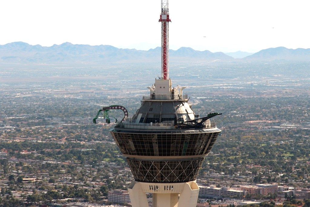 Top of a tall observation tower with thrill rides, overlooking a sprawling desert city and distant mountains