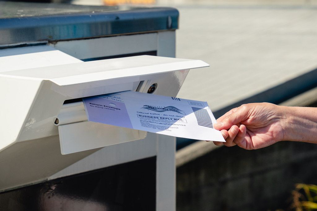 A hand inserts an official ballot return envelope into the slot of a white ballot drop box